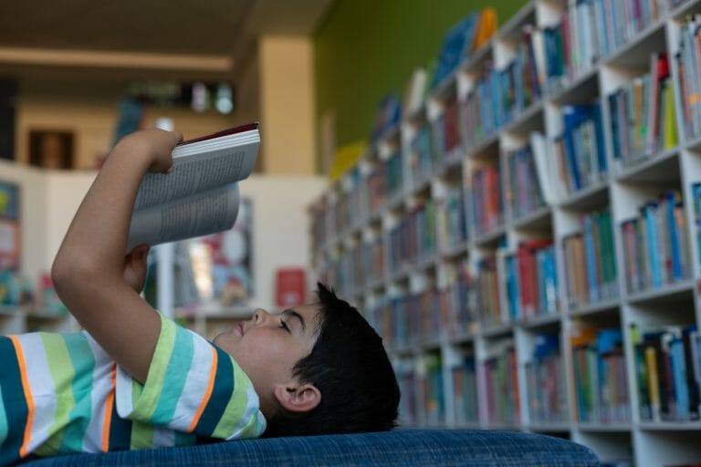 Kobi_Side view of a Caucasian schoolboy lying on the sofa and reading a book in the library at elementary school