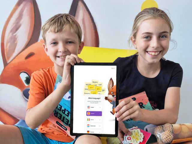 Kids smiling and holding up a tablet with KOBI - Happy Reading app open.